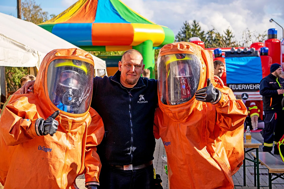 Zwei Besucher in orangen Chemikalienschutzanzügen und ein Feuerwehrmann vor Hüpfburg und Feuerwehrfahrzeugen