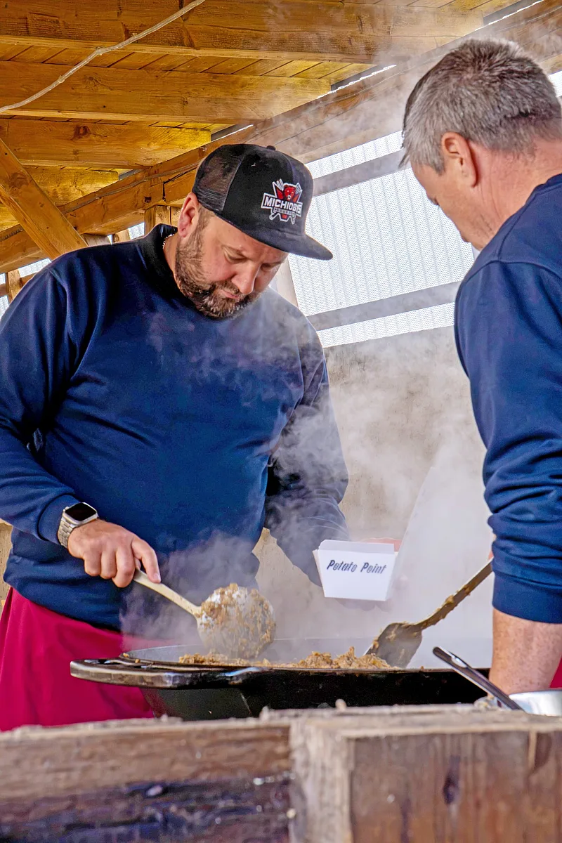 Mann mit Basecap rührt in einer dampfenden Pfanne unter einem Holzdach