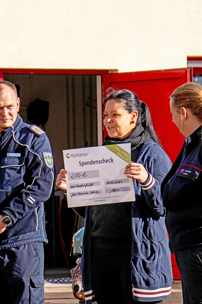 Frau hält einen Spendenscheck, daneben Feuerwehrleute in Uniform